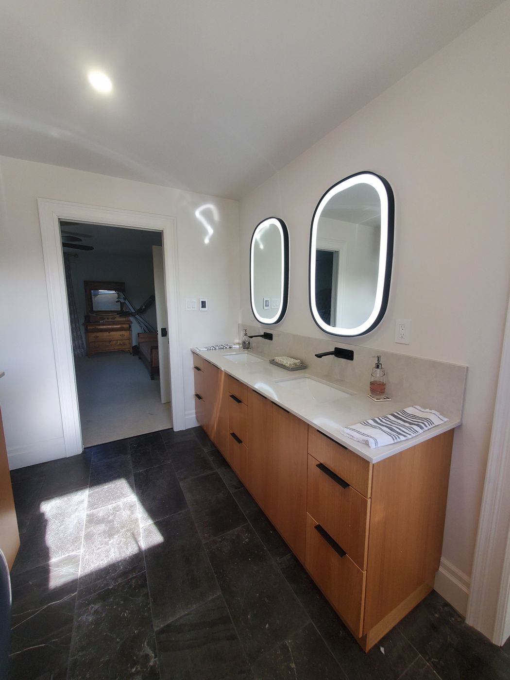 Wood double vanity with lit mirrors on a dark tile floor